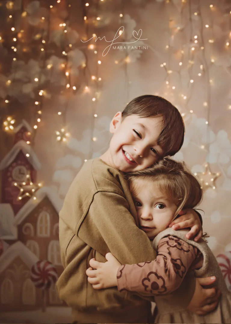 Due fratellini si abbracciano sorridendo davanti a uno sfondo natalizio di casette e lucine dorate