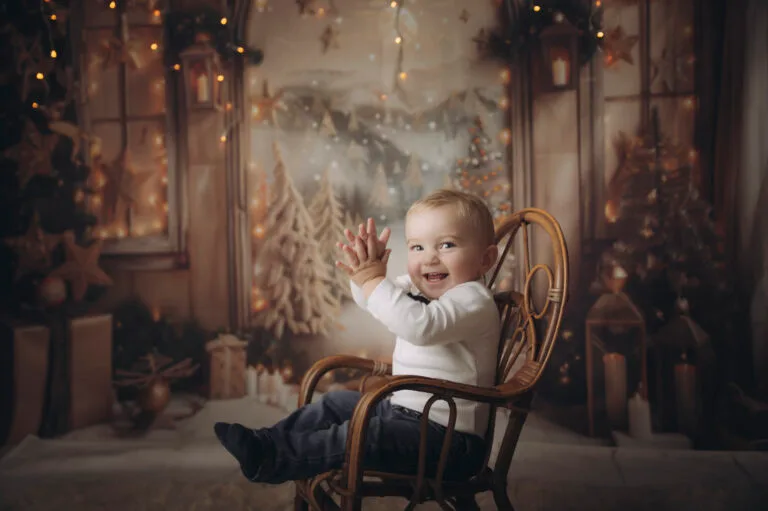 Bambino piccolo che ride e saluta, seduto su una sedia in rattan nel set di Natale con finestra e lanterne